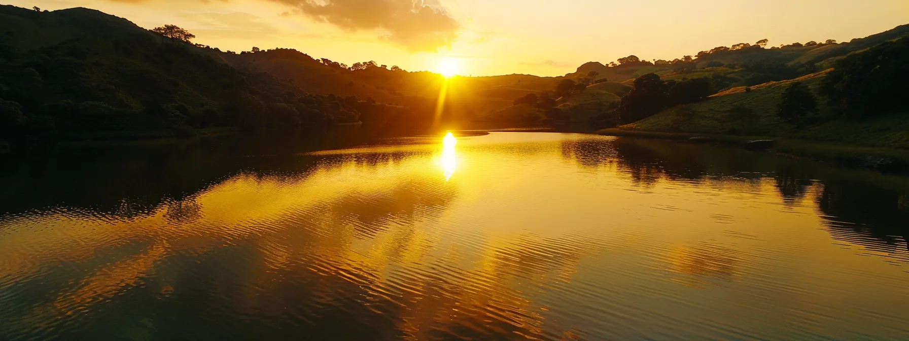 a vibrant sunset casts a warm golden glow over a serene lake, surrounded by lush green hills reflecting in the tranquil water.